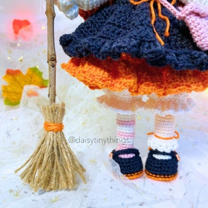 May include: A crocheted witch doll wearing a black and orange dress, white tights, and black shoes. The doll is holding a broom and has a pink bag hanging from her arm. The doll is standing on a white surface with a white and silver background. The text "@daisytinythings" is visible in the image.