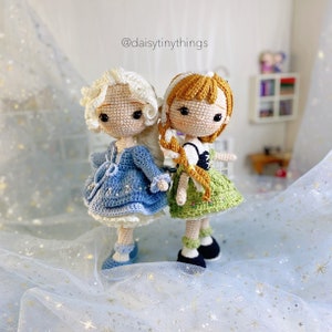 May include: Two crocheted dolls, one with a blue dress and white hair, the other with a green dress and brown hair. The dolls are standing on a white background with gold glitter.