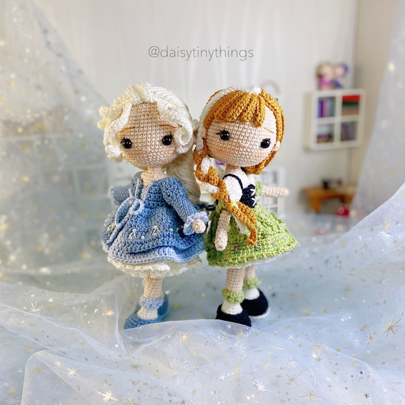 May include: Two crocheted dolls, one with a blue dress and white hair, the other with a green dress and brown hair. The dolls are standing on a white background with gold glitter.