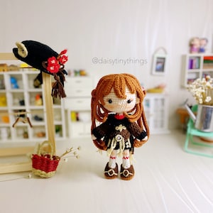 Puede incluir: Una mu&ntilde;eca de crochet con cabello casta&ntilde;o, vestida con un vestido negro y marr&oacute;n, zapatos marrones y una flor roja en su sombrero. La mu&ntilde;eca est&aacute; de pie sobre una superficie blanca. El texto "@daisytinythings" es visible en el fondo.