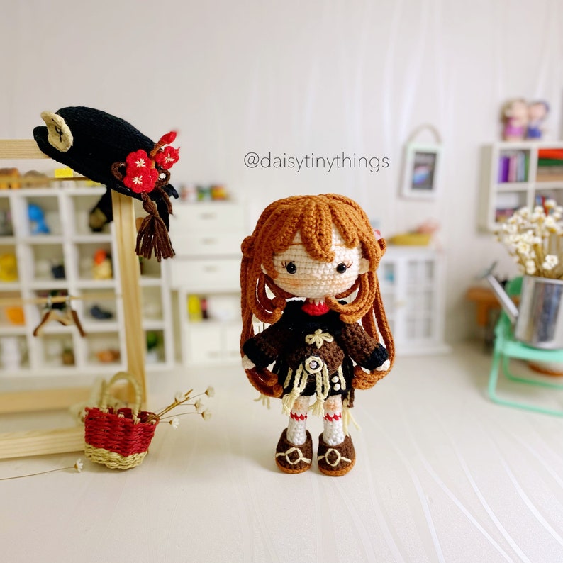 Puede incluir: Una mu&ntilde;eca de crochet con cabello casta&ntilde;o, vestida con un vestido negro y marr&oacute;n, zapatos marrones y una flor roja en su sombrero. La mu&ntilde;eca est&aacute; de pie sobre una superficie blanca. El texto "@daisytinythings" es visible en el fondo.