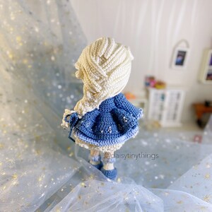 May include: A crocheted doll with long white hair, wearing a blue dress with white snowflake accents. The doll is standing on a blue sparkly surface. The text "@daisytinythings" is visible in the bottom right corner of the image.