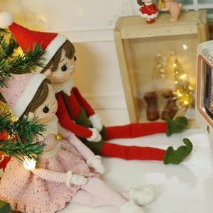 May include: Two crocheted elf dolls, one wearing a red hat and green pants, the other wearing a pink dress, are sitting in front of a miniature television playing a Christmas scene. The television is pink and white with the text "@daisytinythings" on the side.