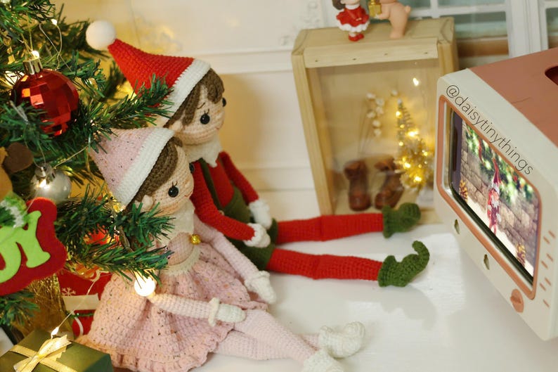 May include: Two crocheted elf dolls, one wearing a red hat and green pants, the other wearing a pink dress, are sitting in front of a miniature television playing a Christmas scene. The television is pink and white with the text "@daisytinythings" on the side.