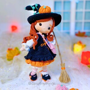 May include: A crocheted witch doll wearing a black and orange dress, a black hat with a pumpkin on top, and holding a broom. The doll has long red hair and brown eyes. The doll is standing on a white surface with a white background.