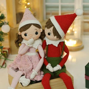 May include: Two crocheted elf dolls, one wearing a pink dress and the other wearing a red and green outfit, sitting on a wooden box. The dolls have brown hair and are holding their hands together. The text "@daisytinythings" is visible on the box.