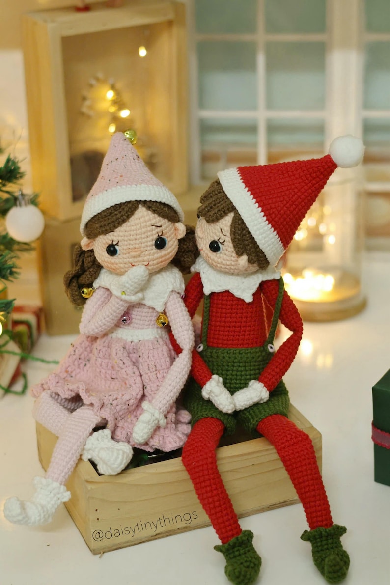 May include: Two crocheted elf dolls, one wearing a pink dress and the other wearing a red and green outfit, sitting on a wooden box. The dolls have brown hair and are holding their hands together. The text "@daisytinythings" is visible on the box.