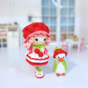 May include: A crocheted doll with pink hair, a red hat, and a white dress with red accents. The doll is holding the hand of a crocheted duck with a red strawberry on its head. The doll is wearing green shoes with white accents. The background is a blurry image of a dollhouse.