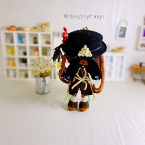 Puede incluir: Una mu&ntilde;eca de crochet con un sombrero negro con un dise&ntilde;o de tri&aacute;ngulo blanco. La mu&ntilde;eca tiene el pelo marr&oacute;n, un atuendo marr&oacute;n y blanco y zapatos marrones. La mu&ntilde;eca est&aacute; de pie sobre una superficie blanca. @daisytinythings