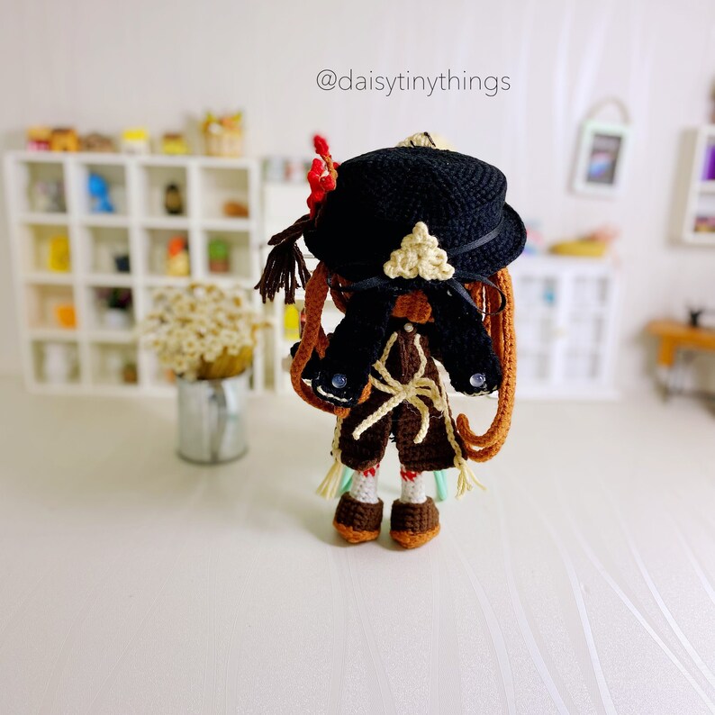 Puede incluir: Una mu&ntilde;eca de crochet con un sombrero negro con un dise&ntilde;o de tri&aacute;ngulo blanco. La mu&ntilde;eca tiene el pelo marr&oacute;n, un atuendo marr&oacute;n y blanco y zapatos marrones. La mu&ntilde;eca est&aacute; de pie sobre una superficie blanca. @daisytinythings