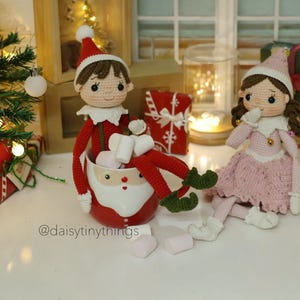 May include: Two crocheted elf dolls, one in a red and green outfit holding a mug filled with marshmallows, the other in a pink dress, sitting on a white surface with a Christmas tree and presents in the background. The text "@daisytinythings" is visible in the bottom left corner of the image.