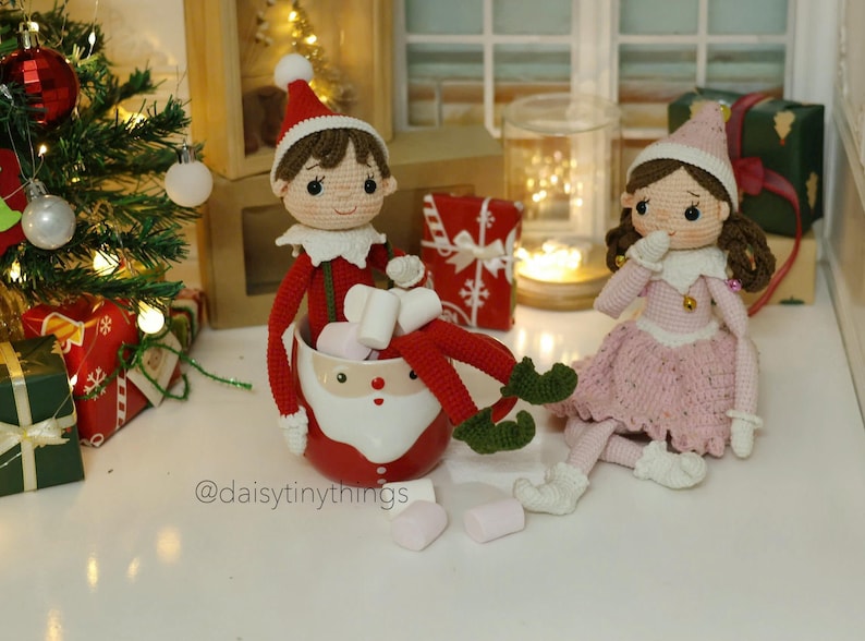 May include: Two crocheted elf dolls, one in a red and green outfit holding a mug filled with marshmallows, the other in a pink dress, sitting on a white surface with a Christmas tree and presents in the background. The text "@daisytinythings" is visible in the bottom left corner of the image.