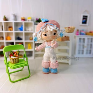 May include: A crocheted doll wearing a pink and blue outfit and a white hat. The doll is holding a small basket of fruit. The doll is standing on a white surface in front of a white shelf with a green chair.