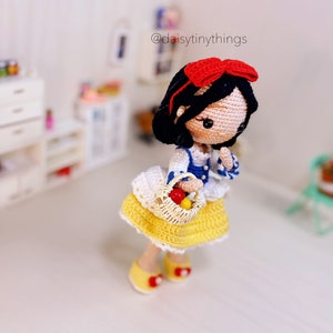 May include: A crocheted Snow White doll with a red bow, blue and white dress, and a basket of red apples. The doll is standing on a white surface.