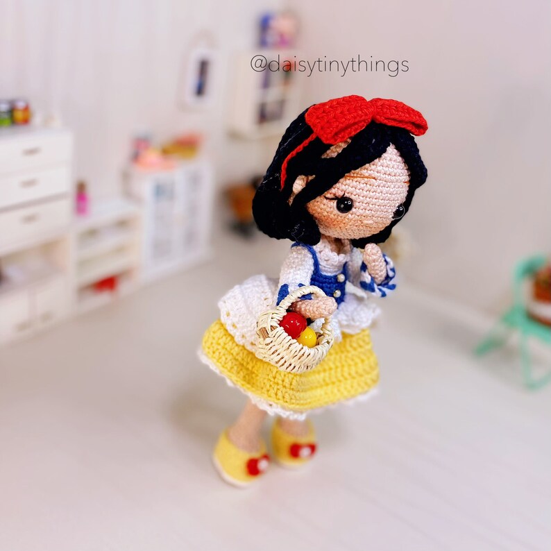May include: A crocheted Snow White doll with a red bow, blue and white dress, and a basket of red apples. The doll is standing on a white surface.