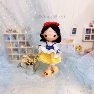 May include: A crocheted Snow White doll with a red bow, yellow dress, blue and white top, and black hair. The doll is standing on a blue tulle fabric with gold stars.