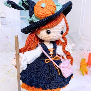 May include: Crocheted witch doll with orange and green hat, a black dress, and a pink purse. The doll is holding a stick and has long red hair. The doll is wearing a white shirt and has a brown belt. The doll is standing on a white surface. @daisytinythings