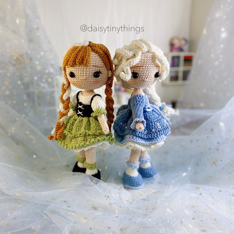 May include: Two crocheted dolls, one with brown hair and a green dress, the other with white hair and a blue dress. The dolls are standing on a white background with a blue and gold glitter pattern. @daisytinythings