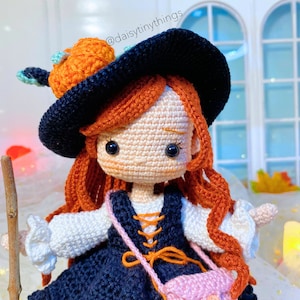 May include: A crocheted witch doll with long red hair wearing a black hat with an orange pumpkin on top. She is wearing a blue and white dress with a pink bag.