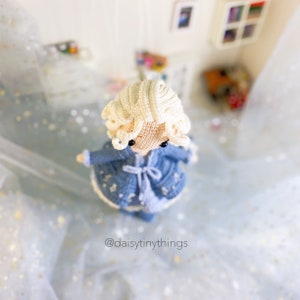 May include: A crocheted doll with blonde hair wearing a blue dress and a white cardigan. The doll is standing on a white background with a light blue and white pattern.