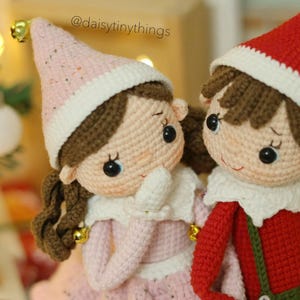 May include: Two crocheted elf dolls, one wearing a pink hat and the other wearing a red hat. Both dolls have brown hair and are smiling.