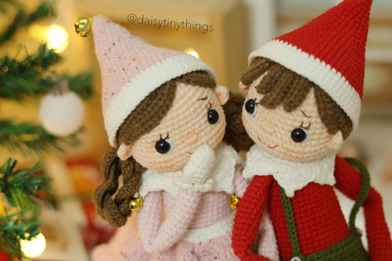 May include: Two crocheted elf dolls, one wearing a pink hat and the other wearing a red hat. Both dolls have brown hair and are smiling.