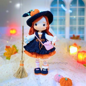 May include: A crocheted witch doll wearing a black hat with an orange pumpkin on top, a blue and orange dress, and pink and white striped stockings. The doll is holding a small pink purse and a broom. The doll is standing on a white surface with two orange pumpkins.
