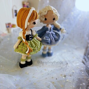 May include: Two crocheted dolls, one with blonde hair and a blue dress, the other with brown hair and a green dress. The dolls are standing on a white background with gold glitter stars. The caption '@daisytinythings' is visible in the bottom right corner of the image.