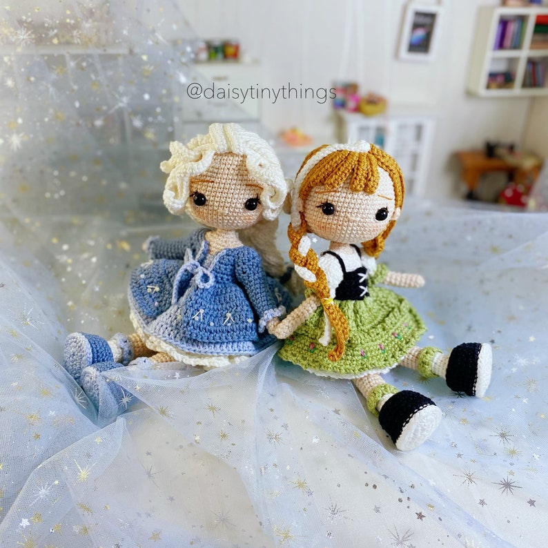 May include: Two crocheted dolls, one with a blue dress and blonde hair, the other with a green dress and brown hair. The dolls are sitting on a blue sparkly fabric with the text "@daisytinythings" in the background.