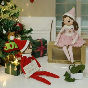 May include: A crocheted elf doll wearing a red and white hat and a red and white outfit. The elf is holding a sign that says "I can't be without you!" The elf is sitting next to a crocheted doll wearing a pink dress and a white hat. The dolls are sitting on a brown box with the text "@daisytinythings" on it.