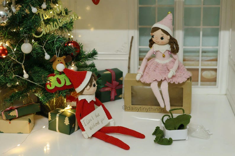 May include: A crocheted elf doll wearing a red and white hat and a red and white outfit. The elf is holding a sign that says "I can't be without you!" The elf is sitting next to a crocheted doll wearing a pink dress and a white hat. The dolls are sitting on a brown box with the text "@daisytinythings" on it.