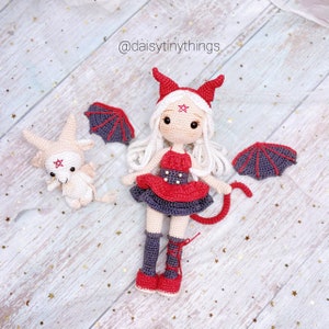 Puede incluir: Una muñeca de crochet vestida de diablo con alas rojas y negras, un vestido rojo y una cola. La muñeca tiene el pelo blanco y una estrella roja en la frente. También hay un animal de crochet más pequeño con pelaje blanco y una estrella roja en la frente en la imagen.