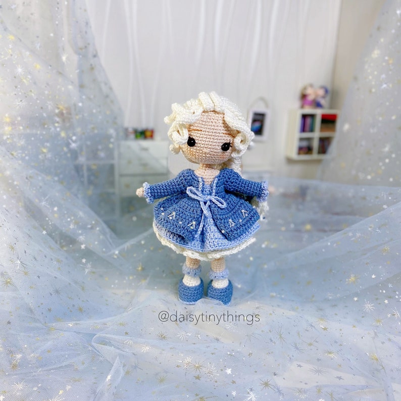 May include: A crocheted doll with long blonde hair wearing a blue dress and a blue cardigan. The doll is standing on a blue and white sparkly fabric.