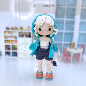 May include: A crocheted doll with white hair, wearing a turquoise jacket, a white tank top, and dark grey shorts. The doll is holding a white and grey bag.