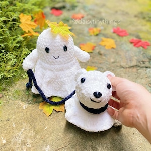 May include: Two white plush toys, a ghost and a bear, are on a path with fall leaves. The ghost has a leaf on its head and is holding a blue leash attached to the bear. The bear has black eyes and a black nose.