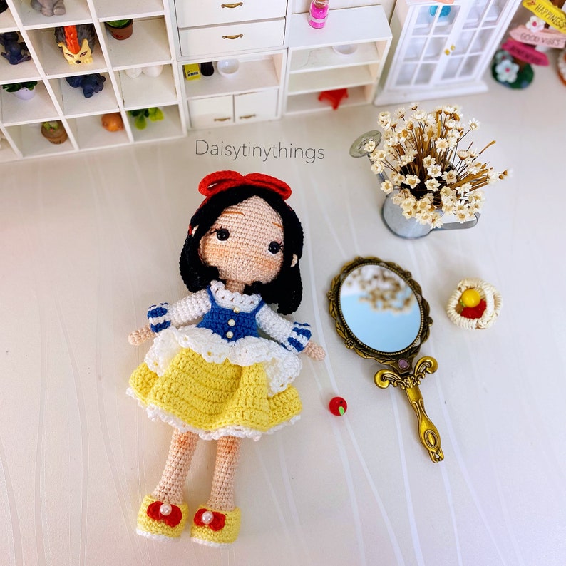 May include: Crocheted Snow White doll with a yellow dress, blue and white top, and red bow. The doll is holding a red apple and a gold mirror with a blue reflection.