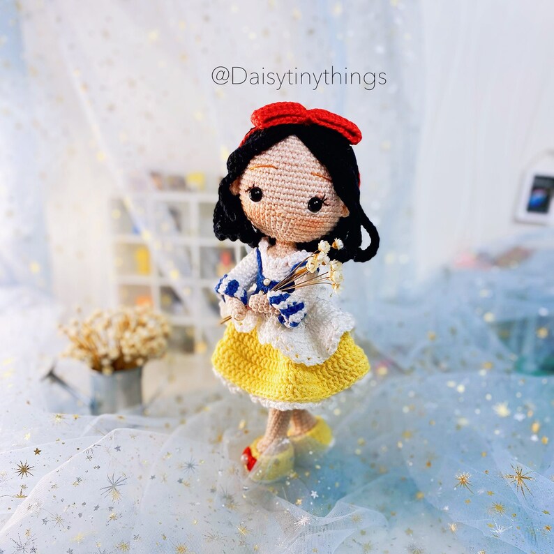 May include: A crocheted doll with black hair, a red bow, and a yellow dress holding a bouquet of white flowers. The doll is standing on a white background with blue stars.