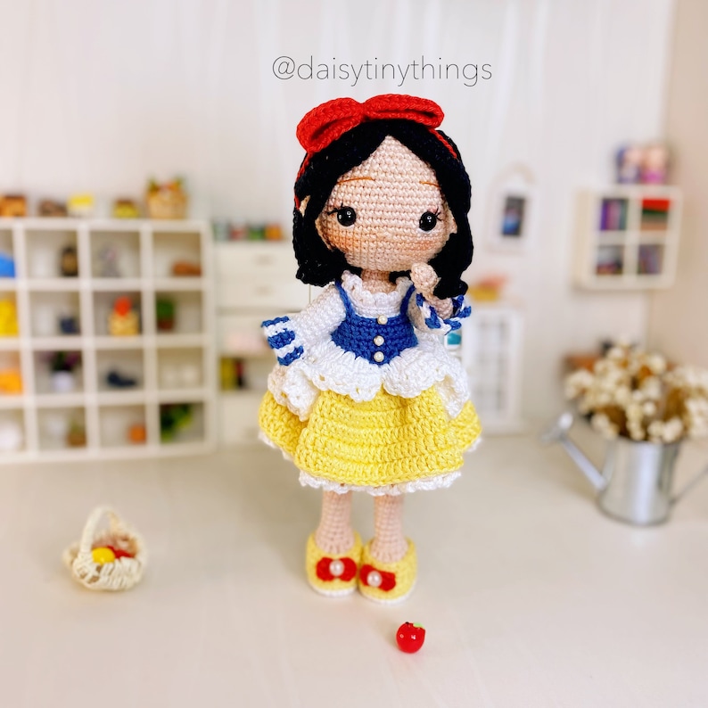 May include: A crocheted Snow White doll with black hair, a red bow, a white and blue dress, and yellow skirt. The doll is holding a red apple in her hand. The doll is standing on a white surface.