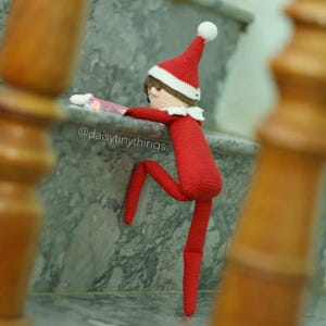 May include: A red crocheted elf doll with a white hat is climbing up a set of stairs. The elf is wearing a red and white striped shirt and has a white pom-pom on its hat. The doll is on a marble staircase with a wooden railing. The Instagram handle @daisytinythings is visible on the stairs.