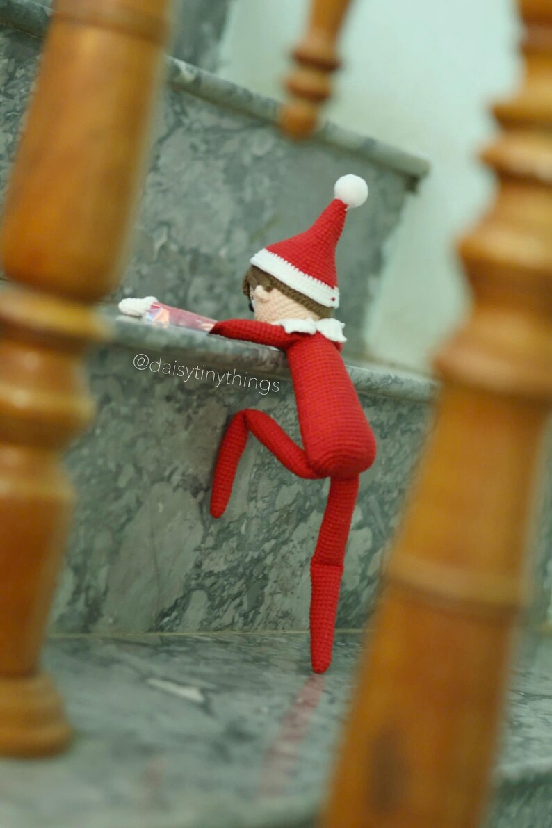 May include: A red crocheted elf doll with a white hat is climbing up a set of stairs. The elf is wearing a red and white striped shirt and has a white pom-pom on its hat. The doll is on a marble staircase with a wooden railing. The Instagram handle @daisytinythings is visible on the stairs.