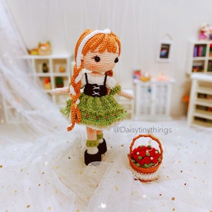 May include: A crocheted doll wearing a green dress with white accents and a white headband. The doll has orange hair and brown eyes. The doll is holding a basket of red flowers. The text "@Daisytinythings" is visible in the image.