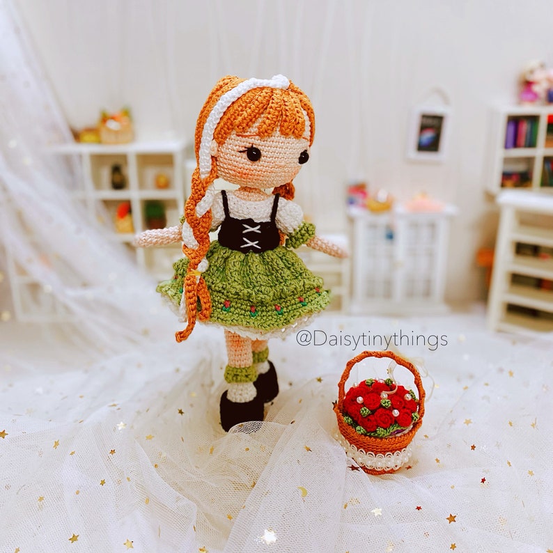 May include: A crocheted doll wearing a green dress with white accents and a white headband. The doll has orange hair and brown eyes. The doll is holding a basket of red flowers. The text "@Daisytinythings" is visible in the image.