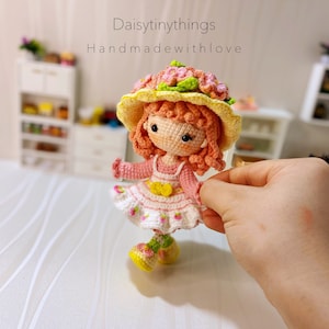 Peut inclure: Une poupée en crochet portant une robe rose avec un nœud jaune, un chapeau jaune avec des fleurs roses et des chaussures vertes. La poupée a les cheveux orange et les yeux bruns. Le texte "Daisytinythings Handmadewithlove" est visible en arrière-plan.