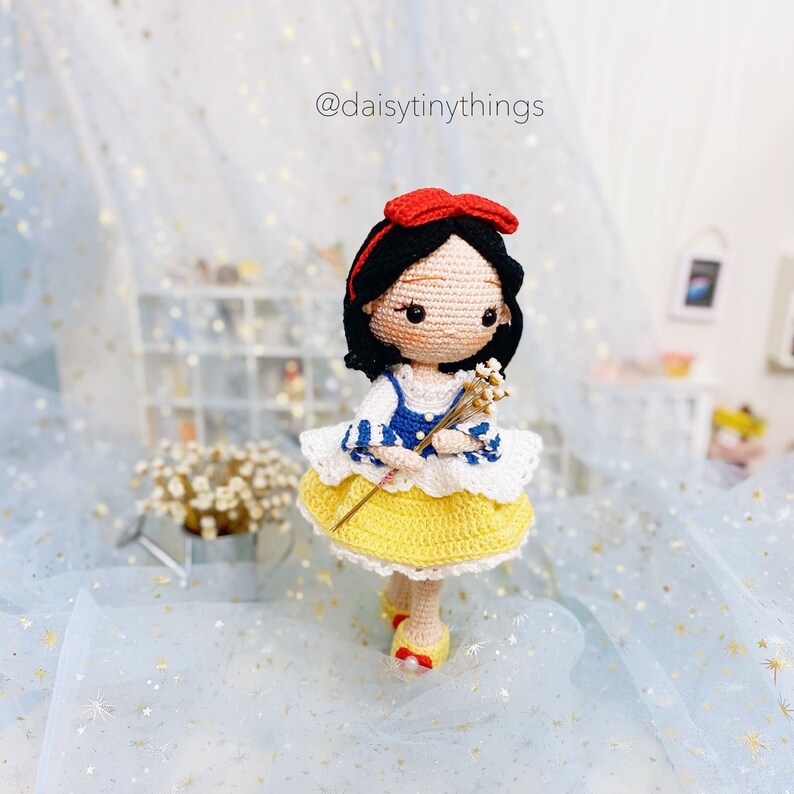 May include: A crocheted Snow White doll with a yellow dress, blue and white striped shirt, and a red bow. She is holding a bouquet of white flowers.