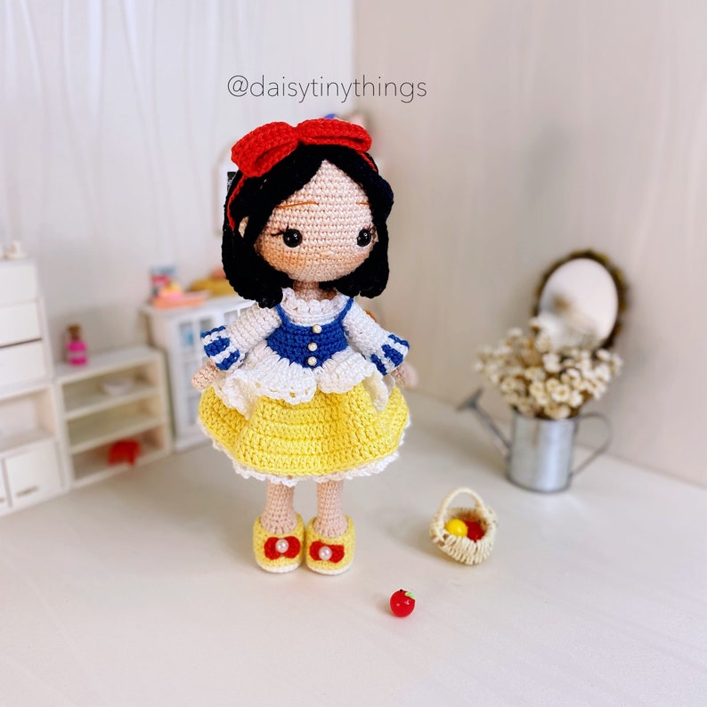 May include: A crocheted Snow White doll with a yellow and white dress, blue vest, and red bow. The doll has black hair and brown eyes. The doll is standing on a white surface with a red apple and a basket of fruit.