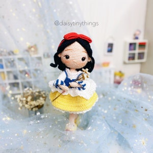May include: A crocheted Snow White doll with a red bow, blue and white striped sleeves, and a yellow skirt. She is holding a white flower bouquet and is standing on a blue tulle fabric with gold stars.