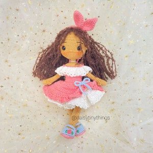 May include: A crocheted doll with brown hair, wearing a pink and white dress with a blue bow. The doll is wearing blue shoes with pink flowers and has a pink bunny ear headband. The doll is sitting on a white background with gold glitter stars.