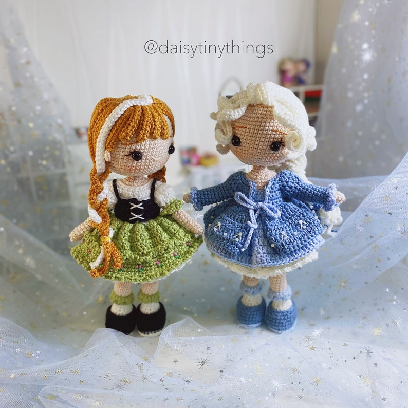 May include: Two crocheted dolls, one with a green dress and brown hair, the other with a blue dress and white hair. The dolls are standing on a white background with a sparkly texture. The text '@daisytinythings' is visible in the top right corner of the image.