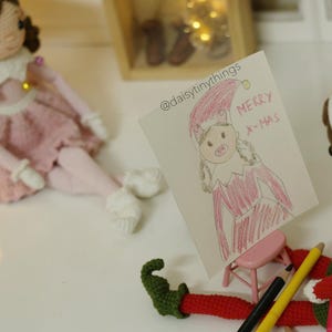 May include: A crocheted elf doll holding a red and white striped pencil. The elf is looking at a drawing of a girl in a red dress with the words "Merry Xmas" on a white card. The card is on a small wooden easel.