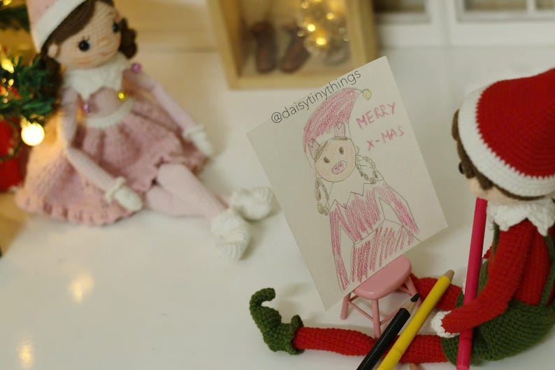 May include: A crocheted elf doll holding a red and white striped pencil. The elf is looking at a drawing of a girl in a red dress with the words "Merry Xmas" on a white card. The card is on a small wooden easel.
